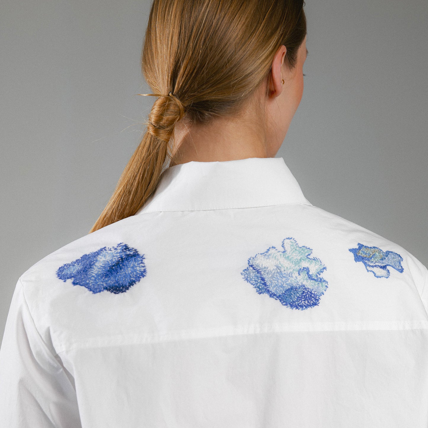 White shirt with blue hand embroidery clouds on the back, worn by a person on a gray background