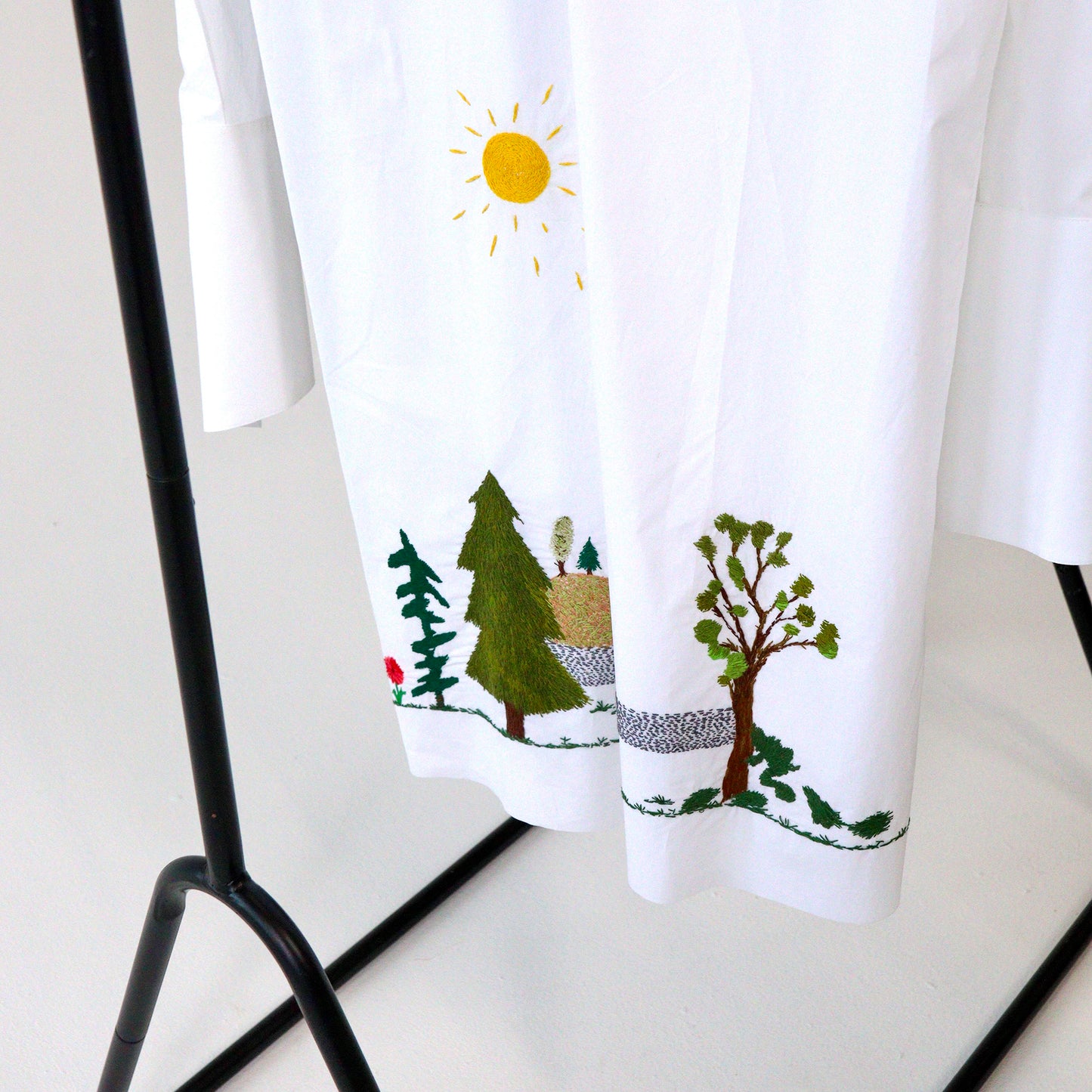 White button up short hanging with nature-themed hand embroidery design featuring trees and a sun, hanging on a black rack.