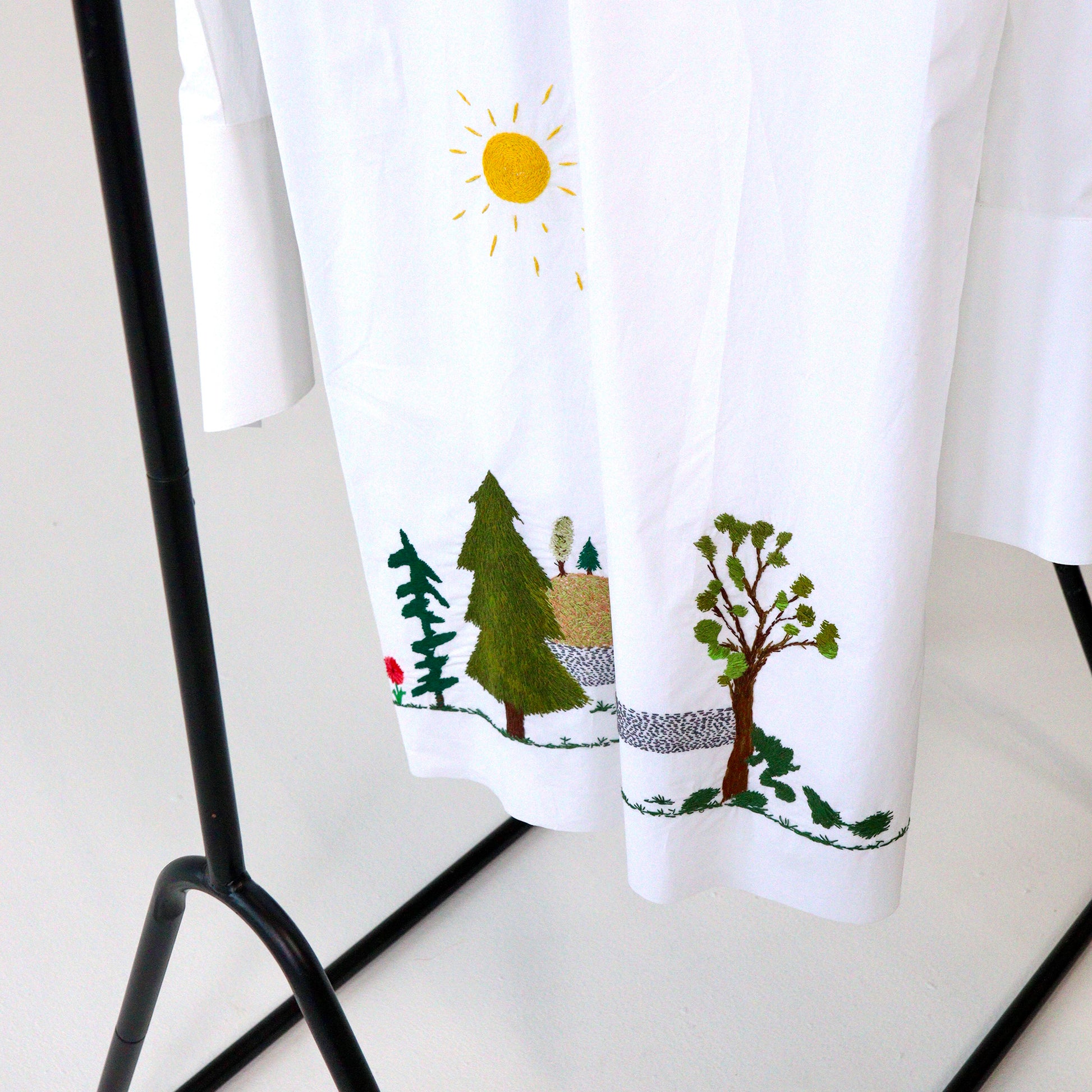 White button up short hanging with nature-themed hand embroidery design featuring trees and a sun, hanging on a black rack.