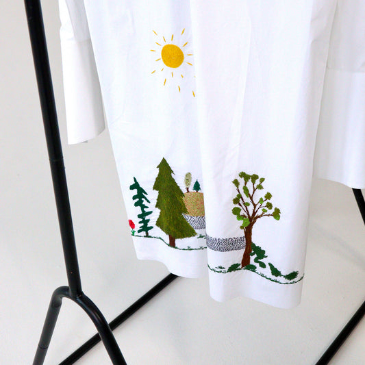 White button up short hanging with nature-themed hand embroidery design featuring trees and a sun, hanging on a black rack.