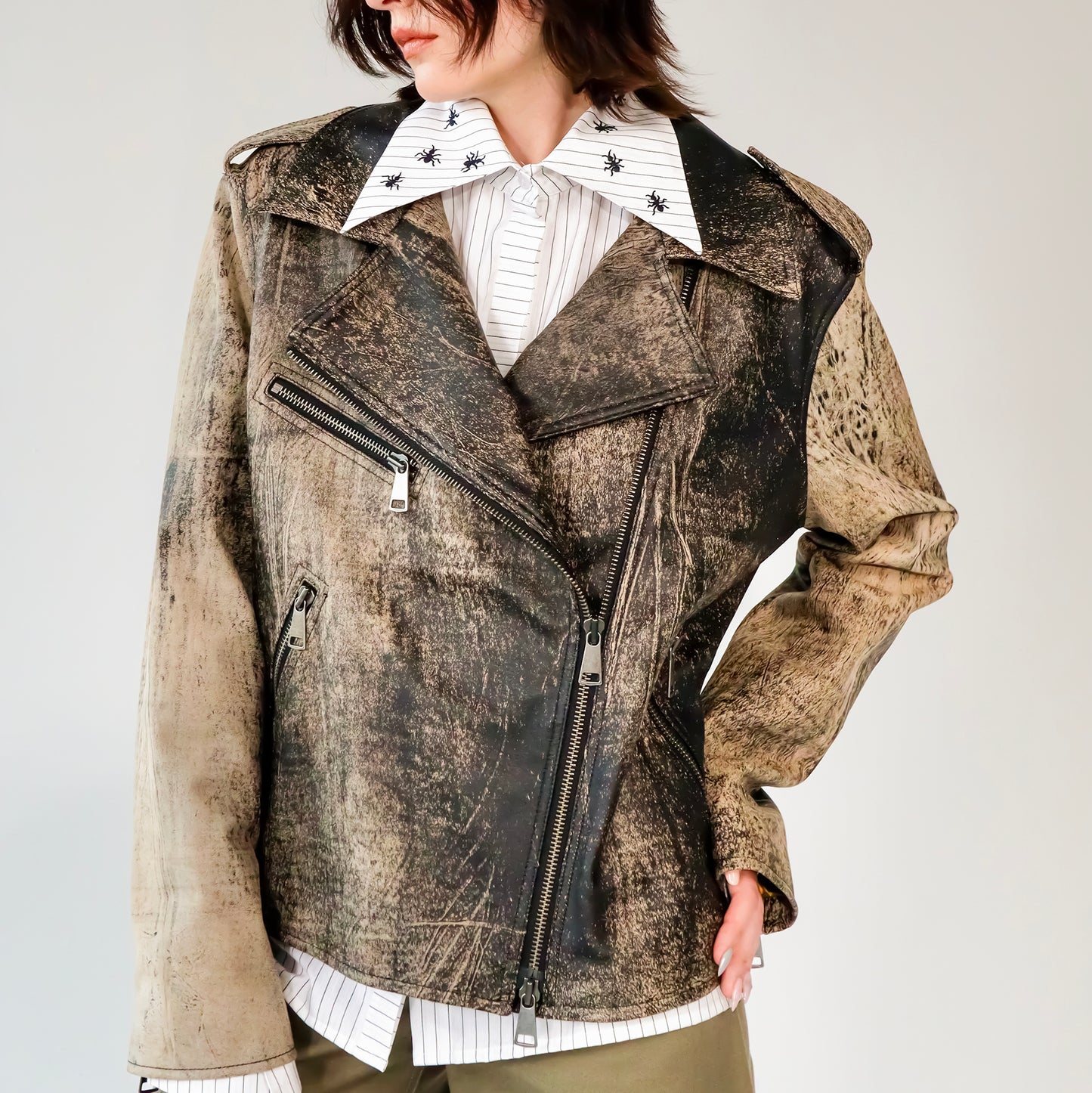 Person wearing a patterned black and brown soft lambskin leather aviator style jacket over a white shirt with black embroidered ant designs on a light gray background. made by mucina studio in latvia