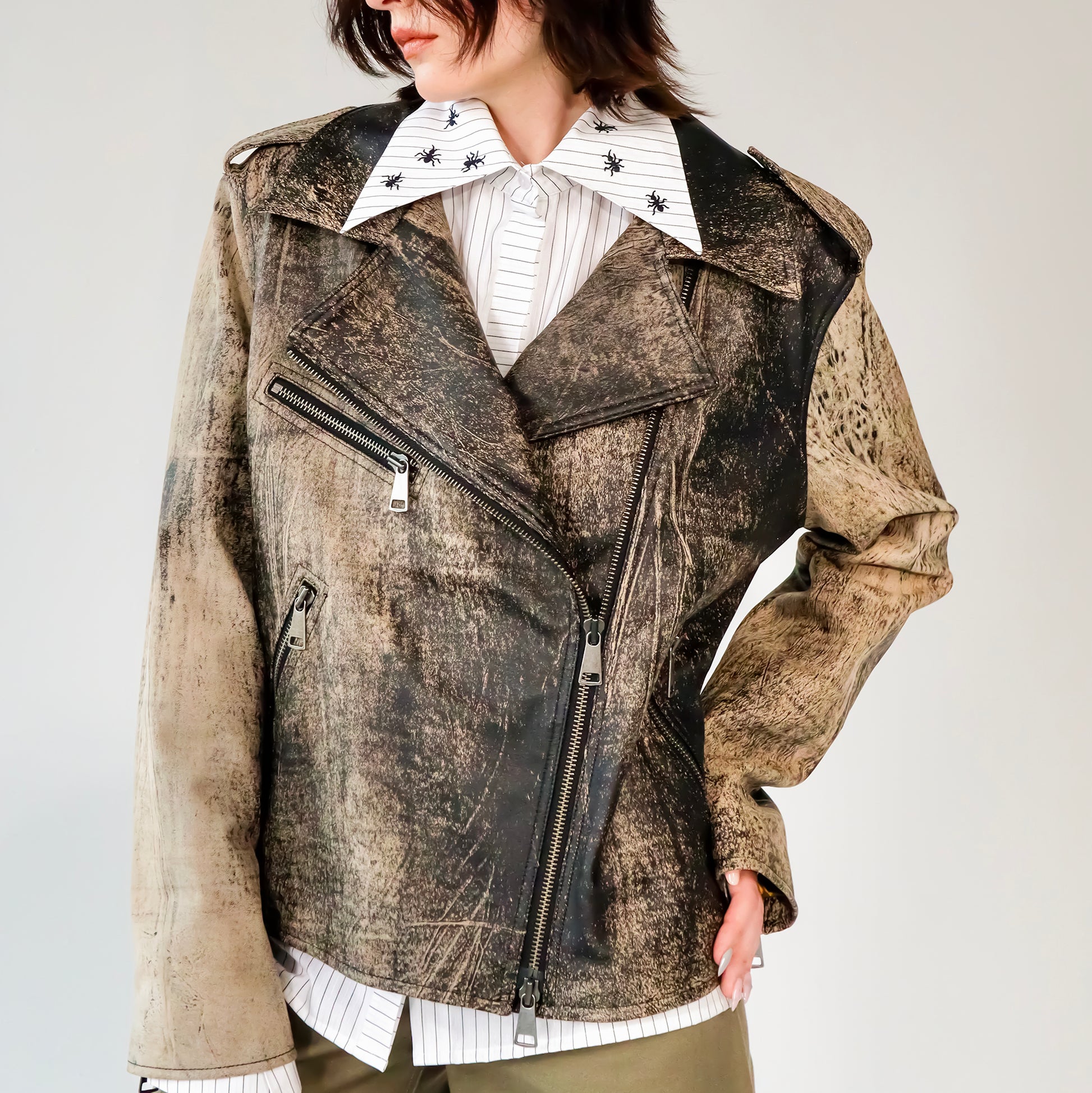 Person wearing a patterned black and brown soft lambskin leather aviator style jacket over a white shirt with black embroidered ant designs on a light gray background. made by mucina studio in latvia