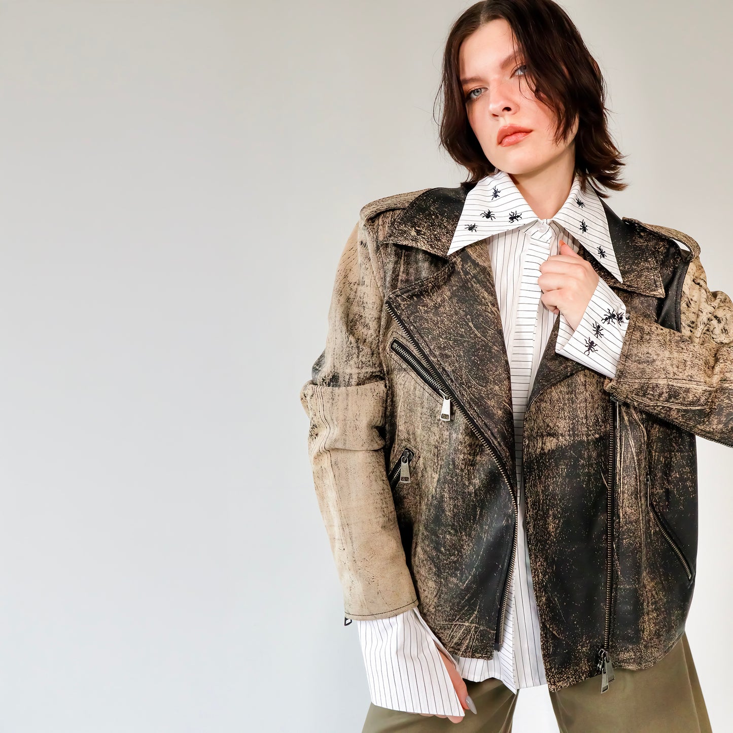 Person wearing a patterned black and brown soft lambskin leather aviator style jacket over a white shirt with black embroidered ant designs on a light gray background. made by mucina studio in latvia