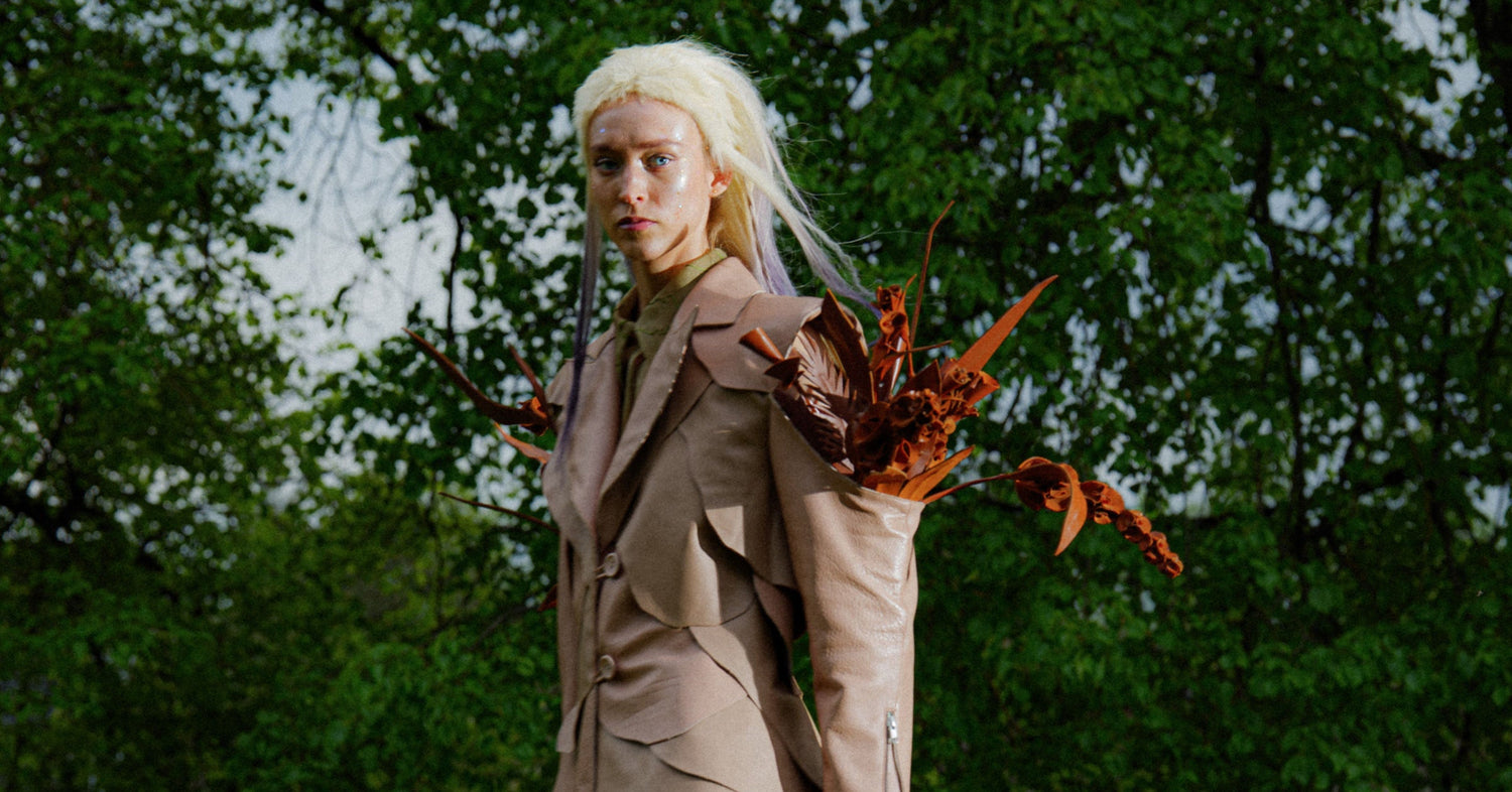 Blonde woman in a wig in a beige leather blazer with floral accessories made from leather against a green forest background