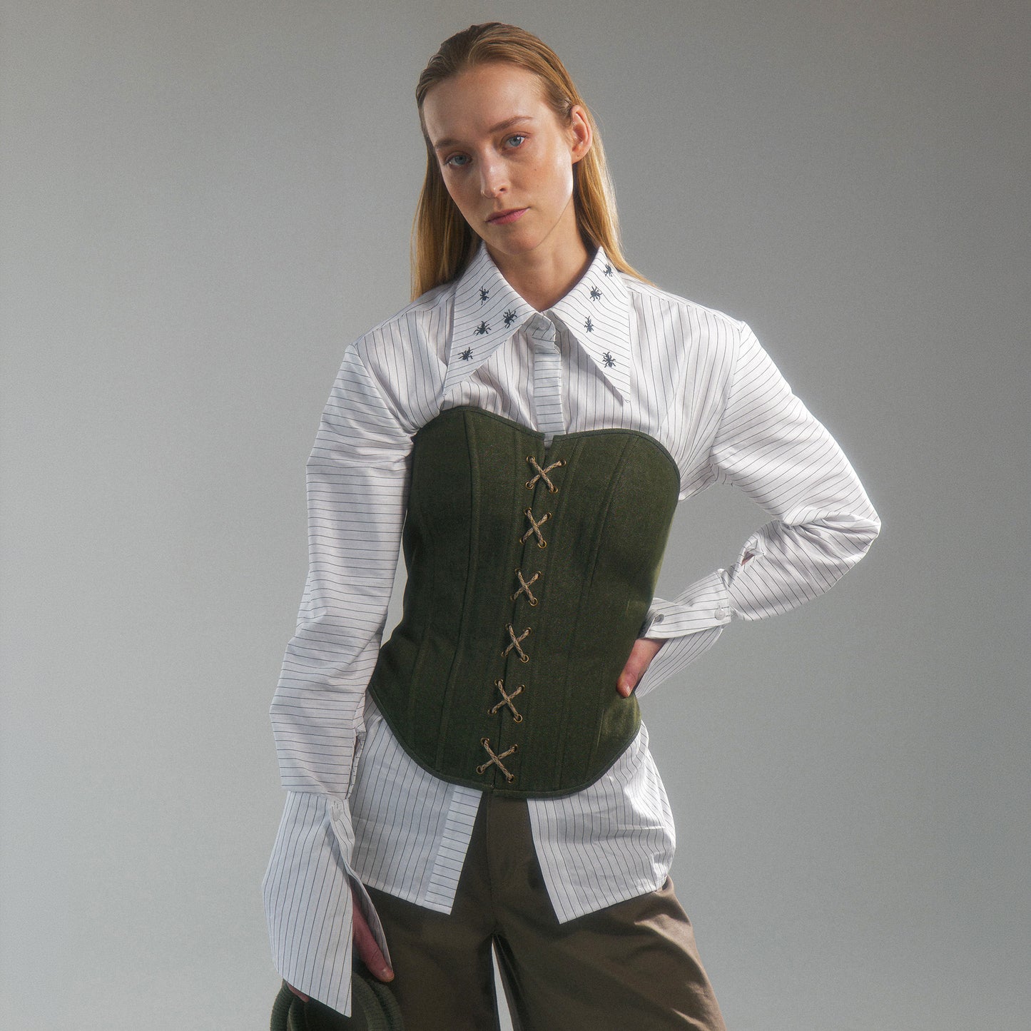 Woman wearing a white shirt with hand embroidered  ant illustrations under a green front lace up corset and khaki wide leg trousers against a gray background