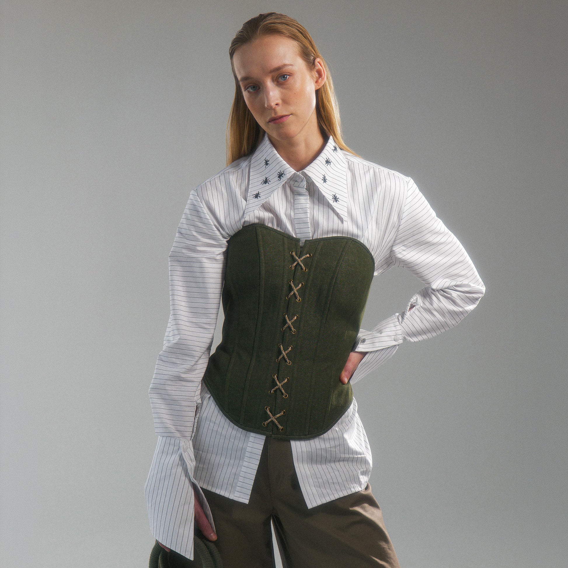Woman wearing a white shirt with hand embroidered  ant illustrations under a green front lace up corset and khaki wide leg trousers against a gray background