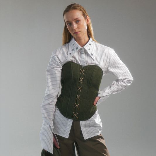 Woman wearing a white shirt with hand embroidered  ant illustrations under a green front lace up corset and khaki wide leg trousers against a gray background