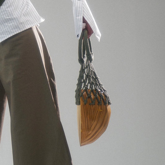 Person holding a half circle wood bag with woven macrame handle next to a person wearing cotton front lace up corset over a button up top and wide leg trousers