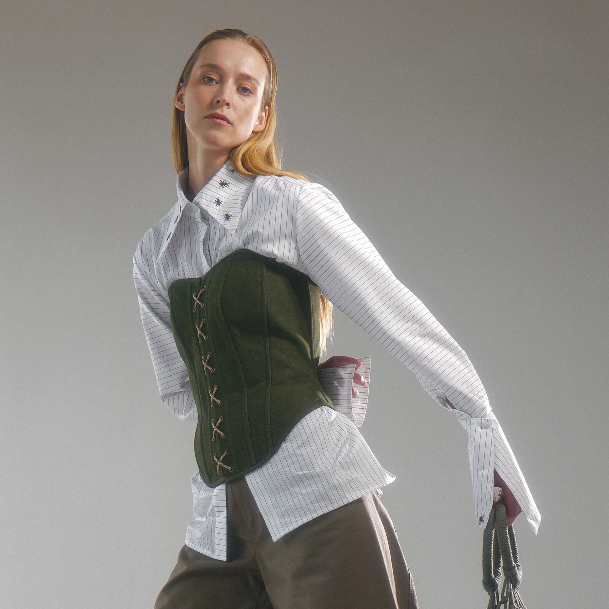 Woman wearing a white shirt with hand embroidered  ant illustrations under a green front lace up corset and khaki wide leg trousers against a gray background