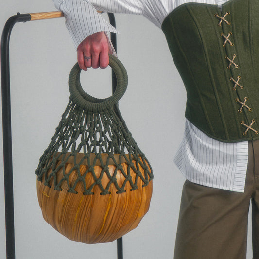 Person holding a half circular wood bag with woven macrame handle next to a person wearing cotton front lace up corset over a button up top and wide leg trousers