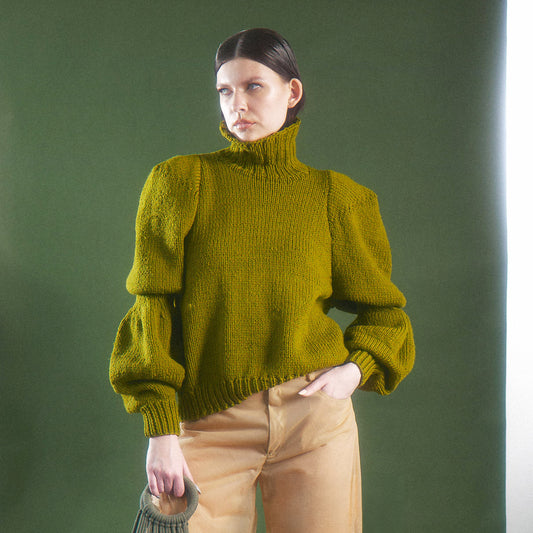 Woman wearing a green hand knitted sweater that has sculptural double-puff sleeves: one puff at the upper arm and another at the forearm, separated by a fitted section. The sleeves taper into long ribbed cuffs, giving the chunky knit a dramatic, voluminous shape. puffed sleeves against a green background