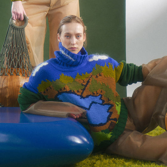 woman sitting in a hand knit merino wool sweater with nature landscape motif represented on the front in an organza silk jumpsuit next to another model holding a wood statement circle bag with macrame details