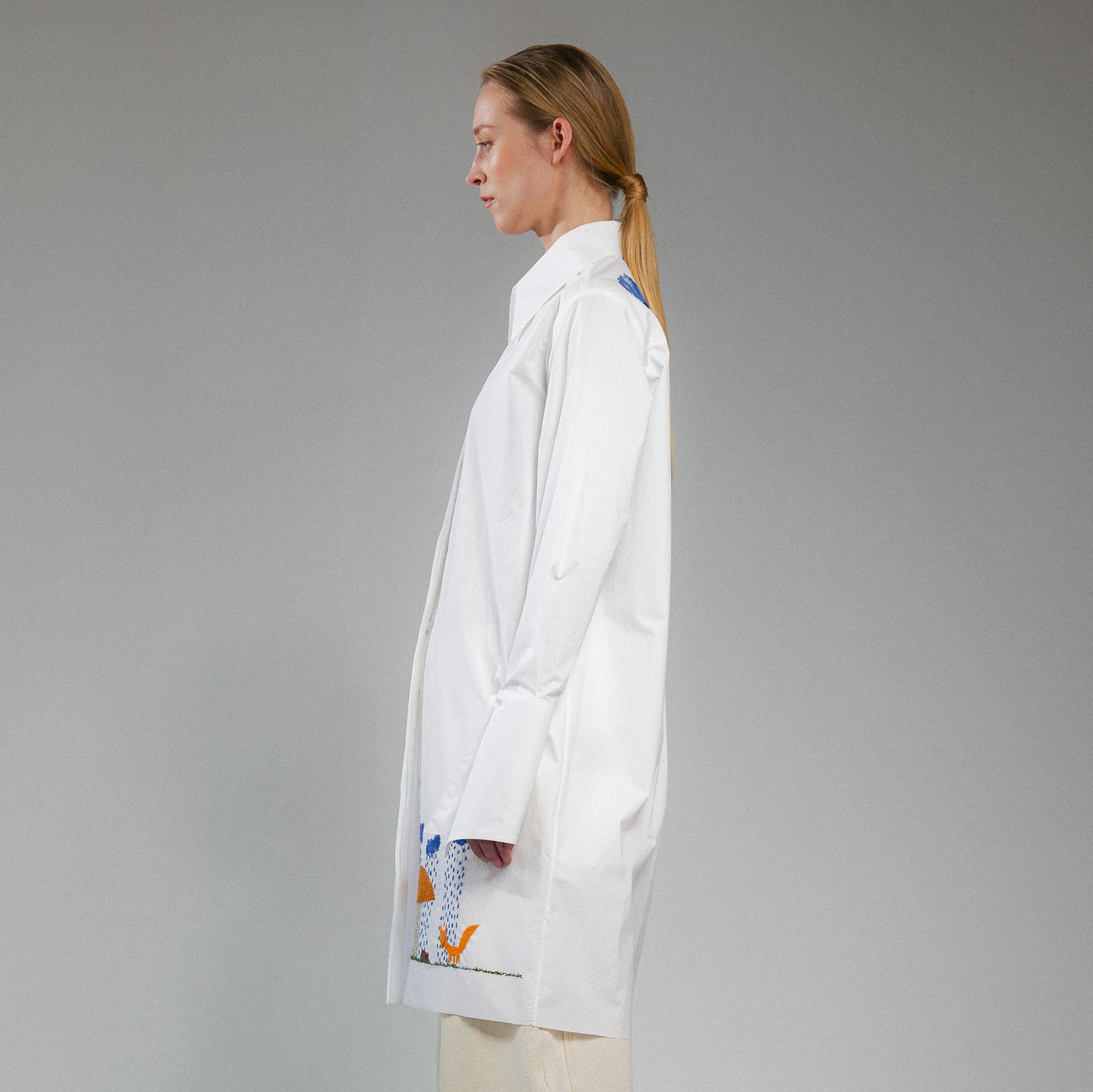 Person wearing a white button up long shirt with hand embroidered designs on a gray background