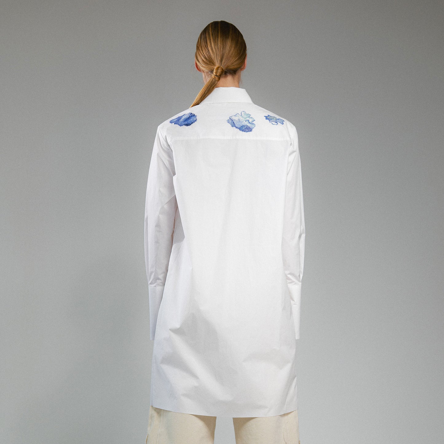 Person wearing a white button up long shirt with hand embroidered designs on a gray background