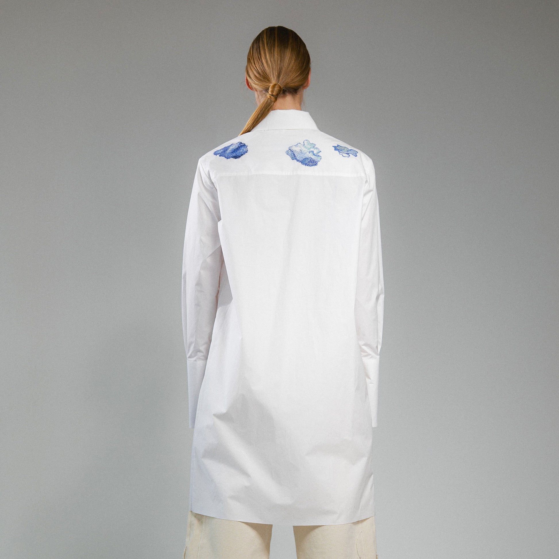 Person wearing a white button up long shirt with hand embroidered designs on a gray background