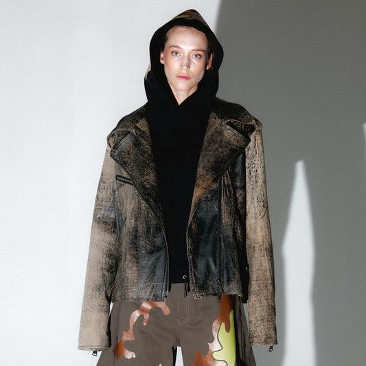 Person wearing a patterned black and brown soft lambskin leather aviator style jacket over a black cropped hoodie with camouflage printed trousers over a gray background. made by mucina studio in latvia