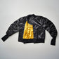 A black bomber jacket with yellow lining, displayed in an unfolded manner against a white background, showcasing the ribbed cuffs and the leather tag on the back.