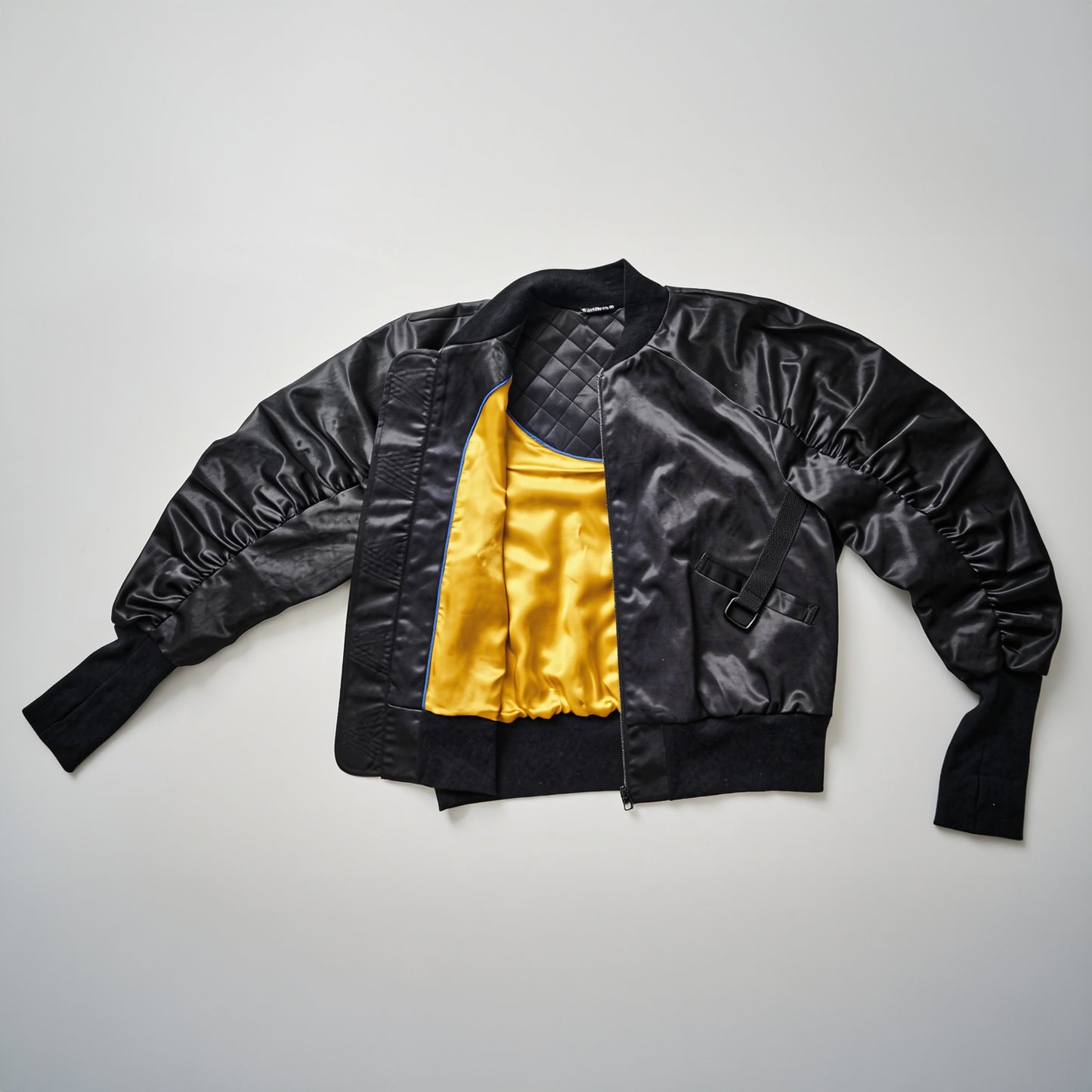 A black bomber jacket with yellow lining, displayed in an unfolded manner against a white background, showcasing the ribbed cuffs and the leather tag on the back.
