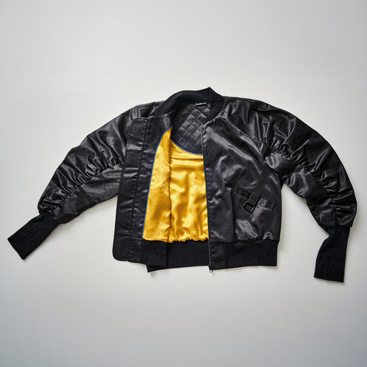 A black bomber jacket with yellow lining, displayed in an unfolded manner against a white background, showcasing the ribbed cuffs and the leather tag on the back.