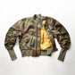 A camouflage bomber jacket laid out flat with the interior lining visible, featuring ribbed cuffs and functional pockets.black streetwear bomber jacket minimalist designer jacket
