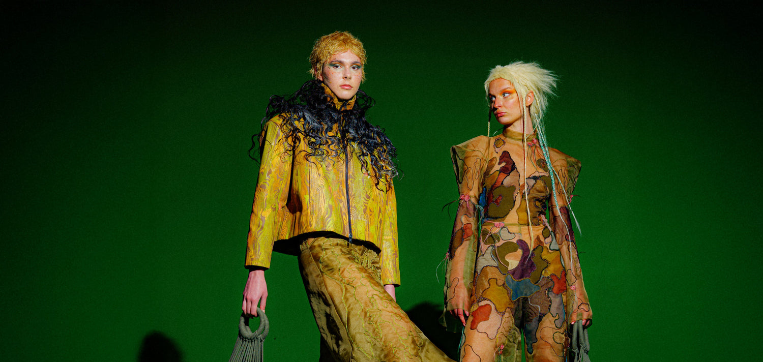 Two models in colorful avant garde hand crafted outfits with wooden sphere bags on a green screen background 